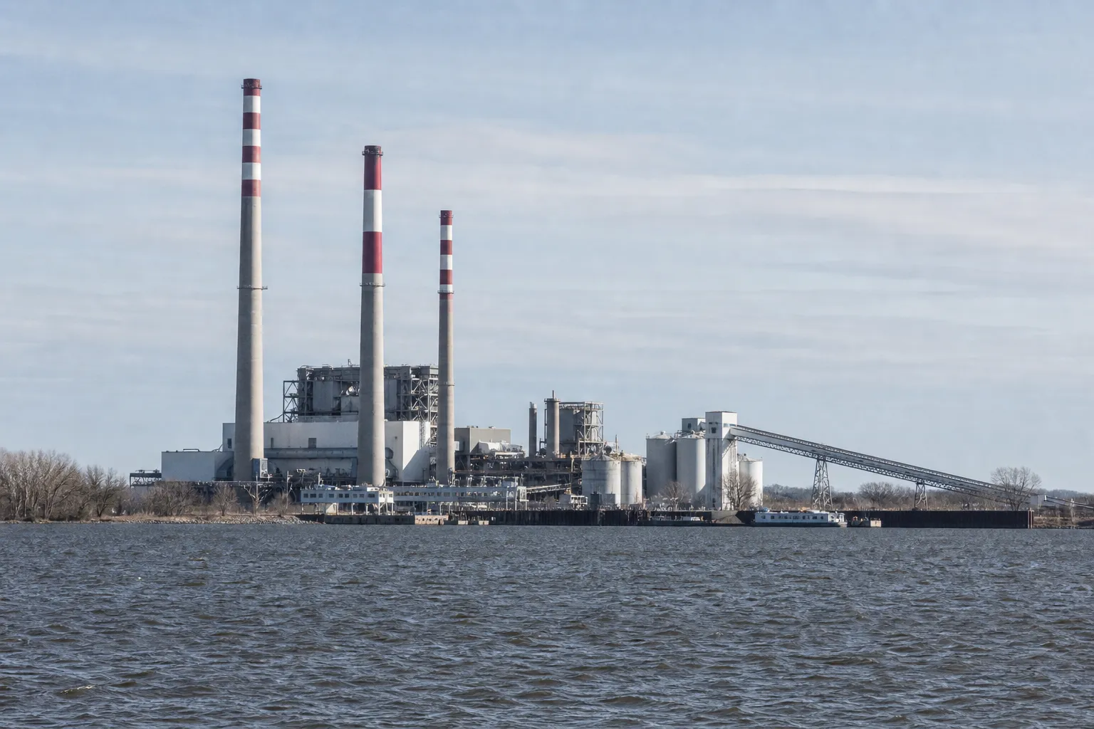 A waterfront thermal power station with red-and-white banded smokestacks, turbine halls, storage tanks, and a conveyor gantry, seen from across the river